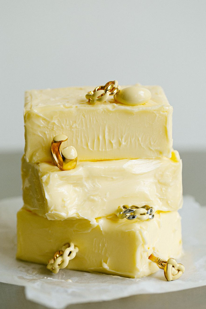 Three stacked rectangular bars of pale yellow butter on parchment paper, each decorated with small gold jewelry pieces, against a soft light gray background. The brand name MISHO is printed at the bottom.